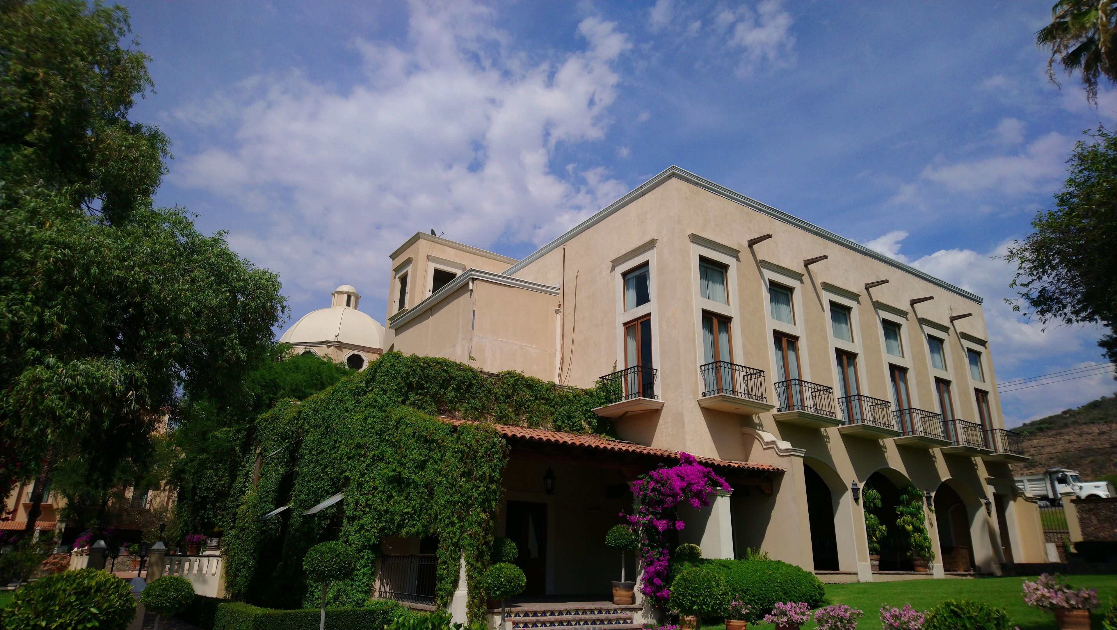 A large building with a dome and multiple balconies, surrounded by lush greenery and a well-maintained garden.