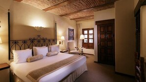 A hotel room with a large bed, wooden ceiling beams, a nightstand, two bedside lamps, a chair, and a door with a window. - Villa Antigua (Tequisquiapan)