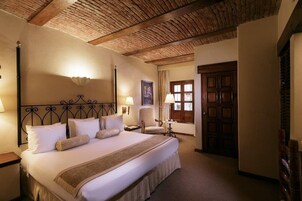 A hotel room with a large bed, wooden ceiling beams, a nightstand, two bedside lamps, a chair, and a door with a window.