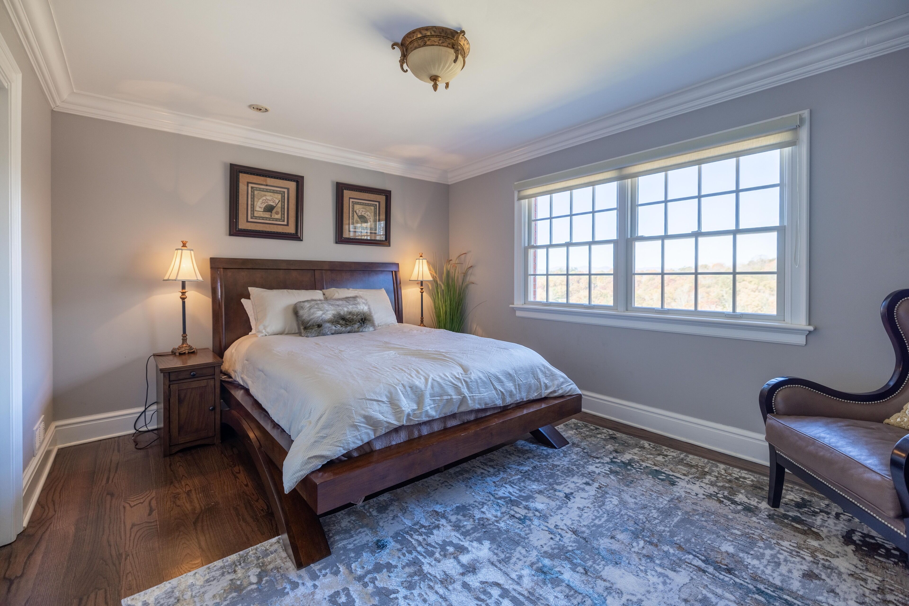 A bedroom with a large bed, two bedside tables, a window with blinds, and a decorative chair.