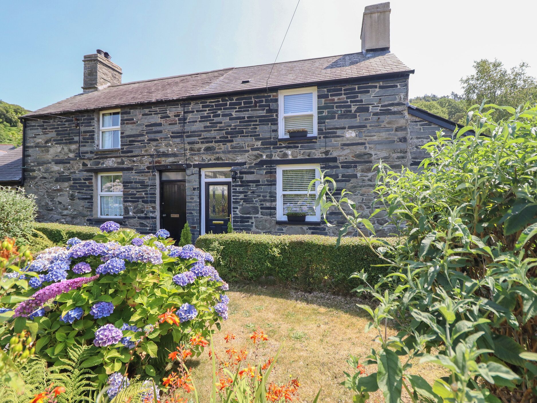 A stone house with a front door, two windows, and a small garden with hydrangeas and other plants.