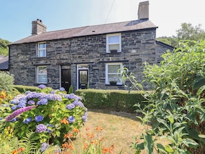 A stone house with a front door, two windows, and a small garden with hydrangeas and other plants. - Arthurs Cottage (Tywyn)