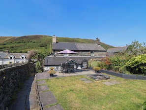 A stone house with a sloped roof, a patio with outdoor furniture, and a garden with various plants. - Arthurs Cottage (Tywyn)