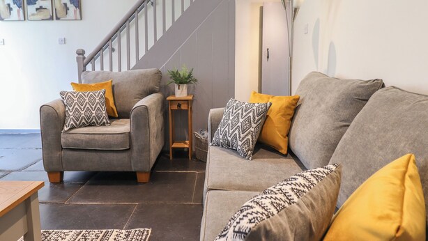 Un salon avec un canapé gris, deux fauteuils et une petite table d’appoint en bois sur laquelle est posée une plante. On aperçoit un escalier au fond.