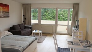 A modern hotel room with a bed, a sofa, a dining table, and a view of greenery through large windows.