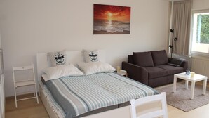 A bedroom with a bed, a sofa, a small table, and a painting of a sunset over the ocean.