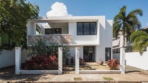 Una casa moderna de dos pisos con techo plano, un balcón y un pequeño jardín con plantas rojas.