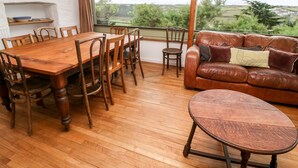 A dining area with a wooden table and chairs, a leather sofa, and a view of the countryside through a large window. - Porth Farm Annexe (Newquay)