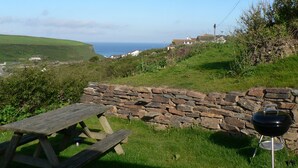 A stone wall, a picnic table, and a barbecue grill in a grassy outdoor area with a view of the sea and houses in the distance. - Porth Farm Annexe (Newquay)