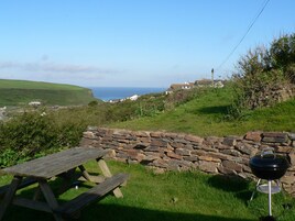 Un muro de piedra, una mesa de picnic y una parrilla en un área verde al aire libre, con vista al mar y casas a lo lejos.
