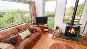 A cozy living room with a leather sofa, a flat-screen TV, a wood-burning stove, and a view of the countryside. - Porth Farm Annexe (Newquay)