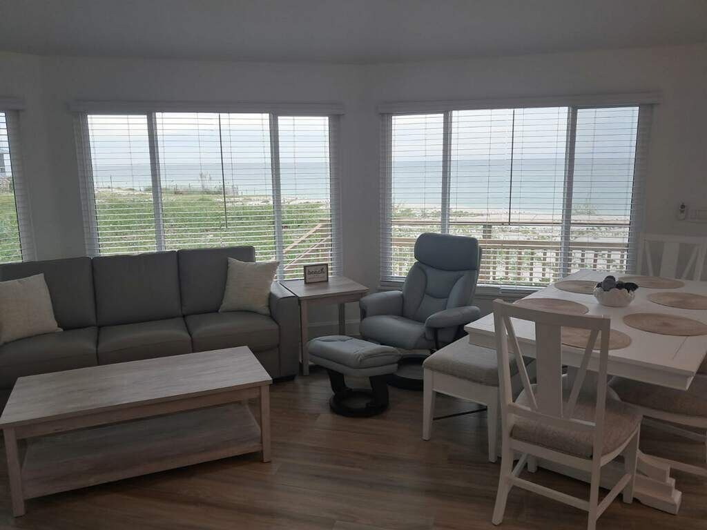 A living room with a sofa, a chair, a dining table, and a view of the sea through large windows.