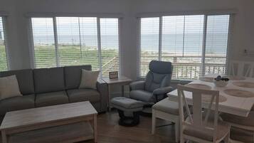 A living room with a sofa, a chair, a dining table, and a view of the sea through large windows.