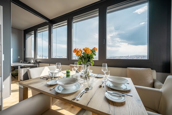 A dining area with a wooden table set for a meal, large windows with a city view, and a sofa in the background.