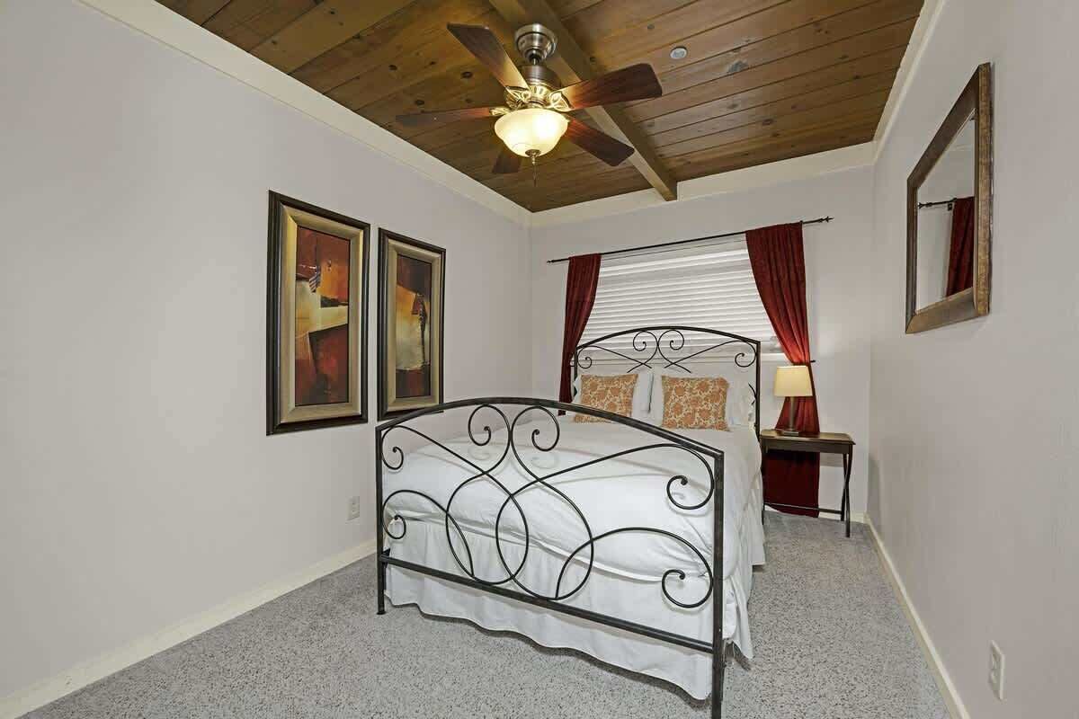 A bedroom with a bed, ceiling fan, two framed pictures, a window with curtains, and a bedside table with a lamp.