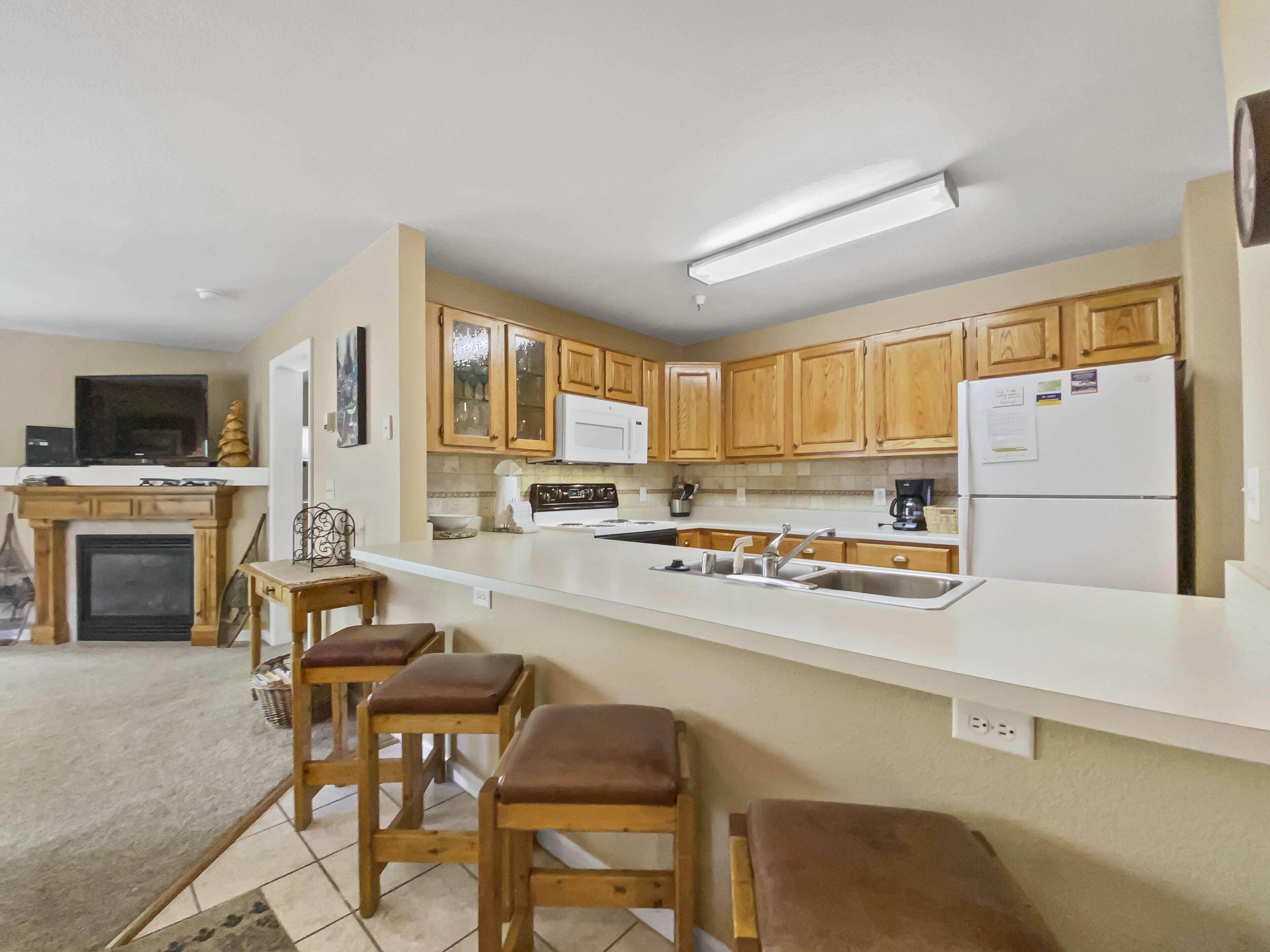 Una cucina moderna con mobili in legno, un piano di lavoro bianco e un piano cottura integrato. Sono presenti tre sgabelli in legno e un camino.