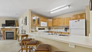 A modern kitchen with wooden cabinets, a white countertop, and a built-in stove. There are three wooden stools and a fireplace.