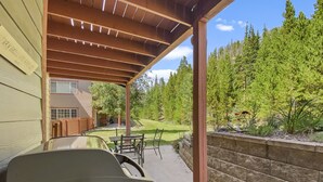 A covered patio area with a barbecue grill, outdoor seating, and a view of a wooded backyard.