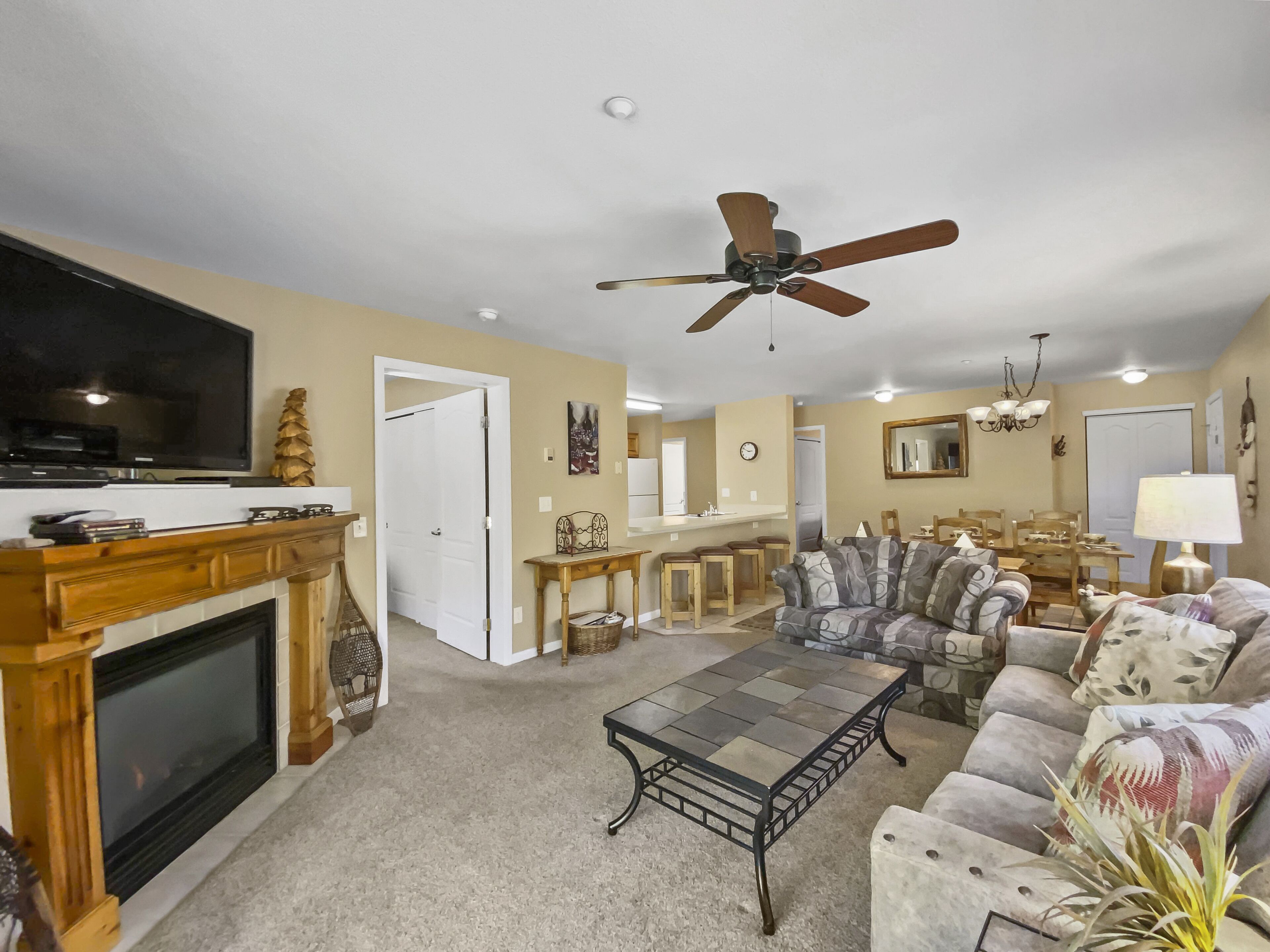 A living room with a fireplace, a ceiling fan, a television, and a dining area with a kitchen in the background.
