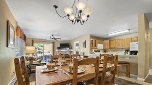 A dining area with a wooden table set for four, surrounded by wooden chairs. A chandelier hangs from the ceiling, and a ceiling fan is visible. The room has wooden cabinets and a television mounted on the wall.
