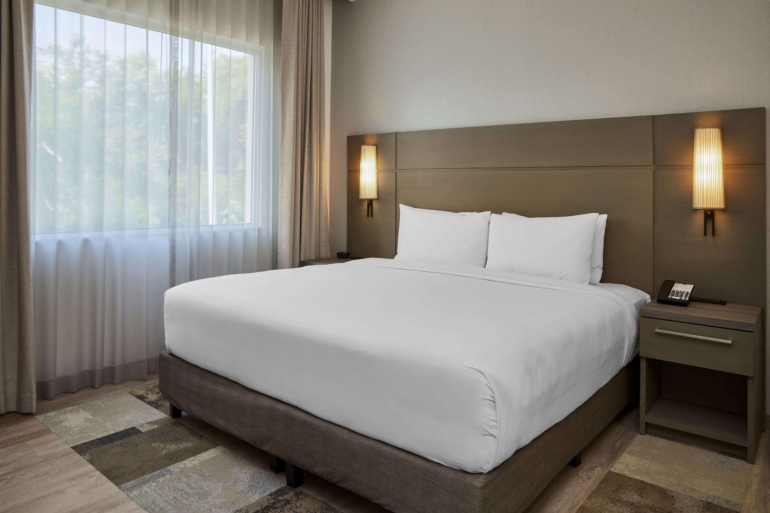 Una cama grande con sábanas blancas, dos mesitas de noche con lámparas y vistas a árboles a través de cortinas traslúcidas.