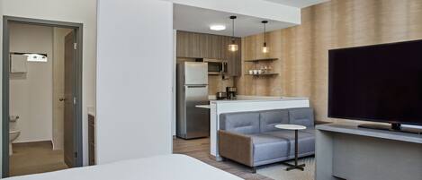 A modern hotel room with a bed, a TV, a kitchenette, and a seating area.