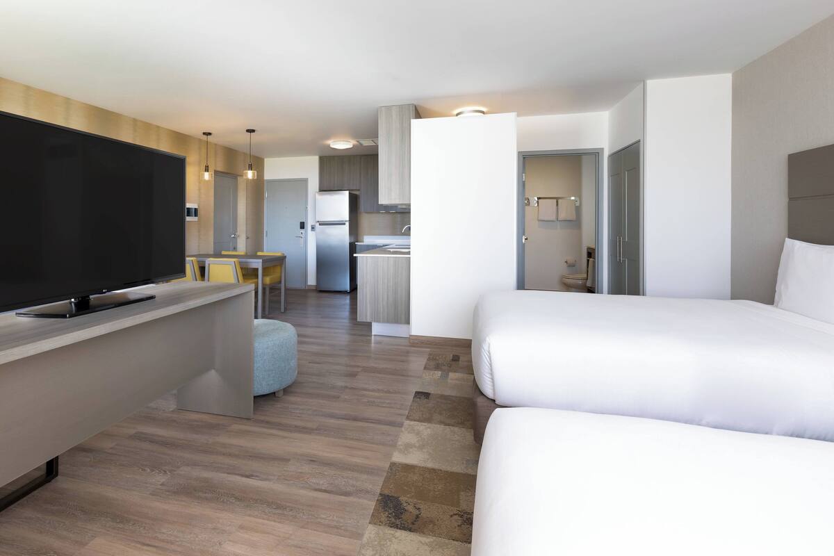 Habitación de hotel moderna con televisor de pantalla plana, sofá, zona de comedor con mesa y sillas, y kitchenette.