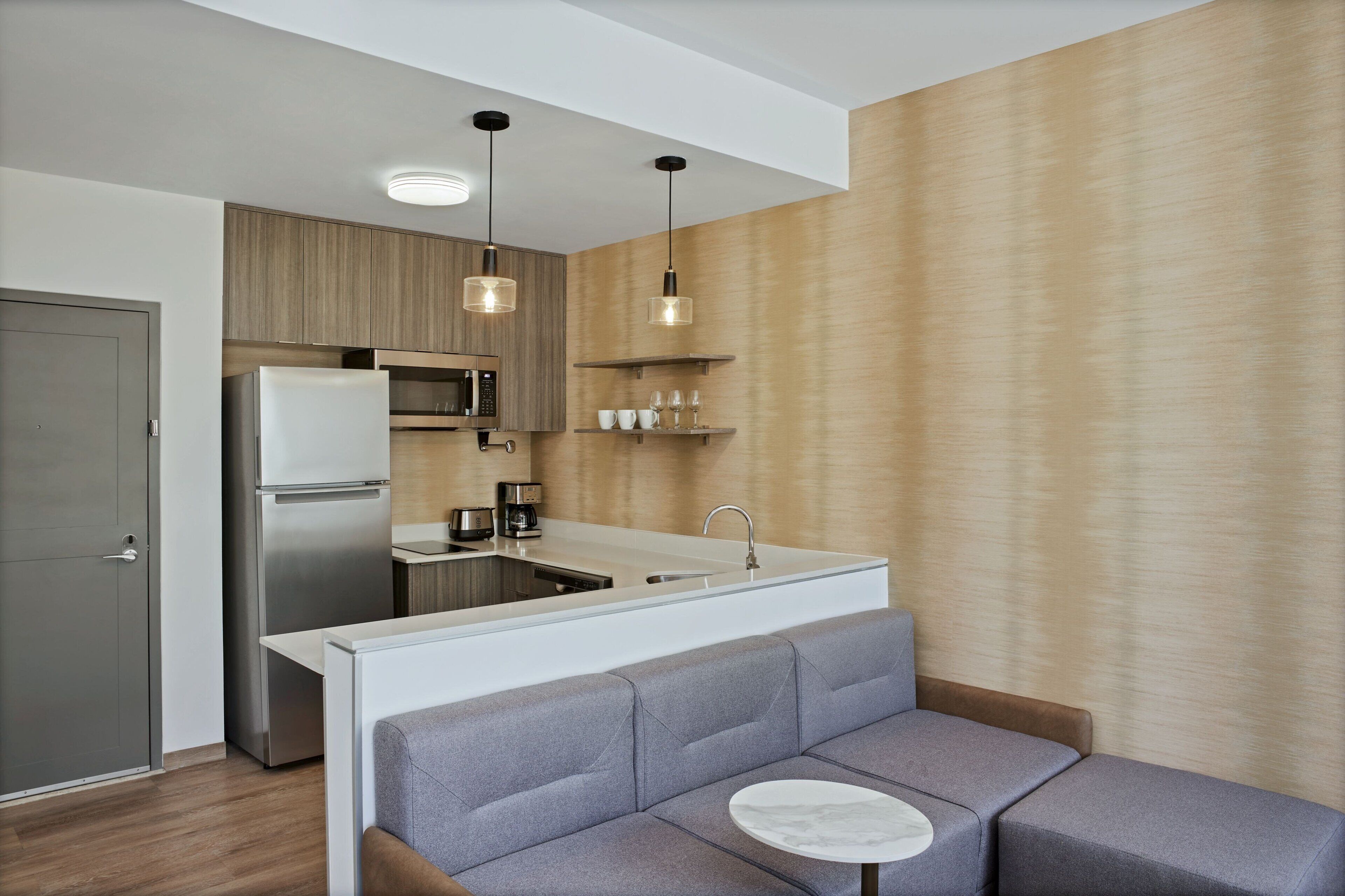 A modern kitchen with a built-in refrigerator, microwave, and sink. There is a gray sofa with a small round table in front.