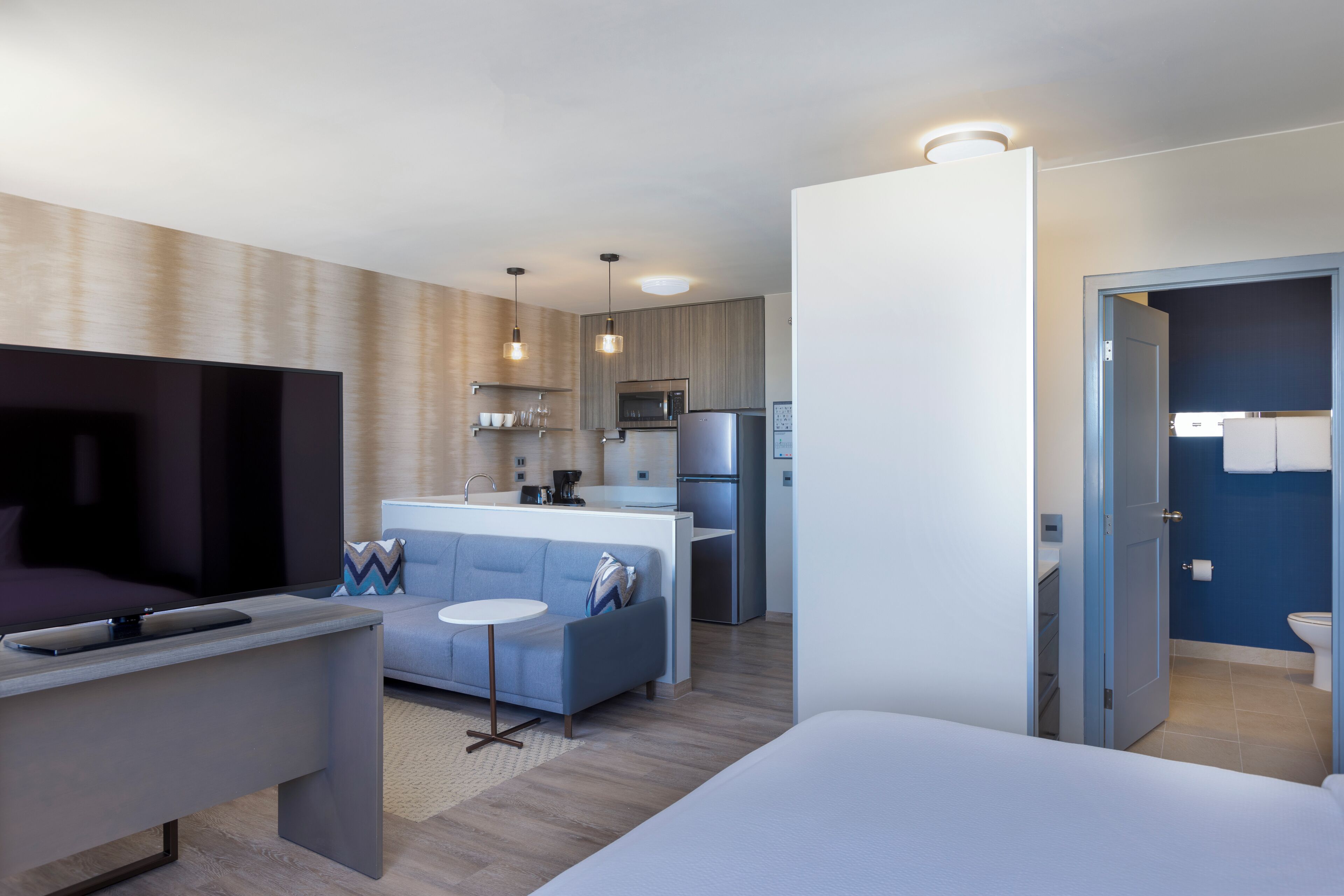 A modern hotel room with a bed, a TV, a sofa, a small table, and a kitchenette.