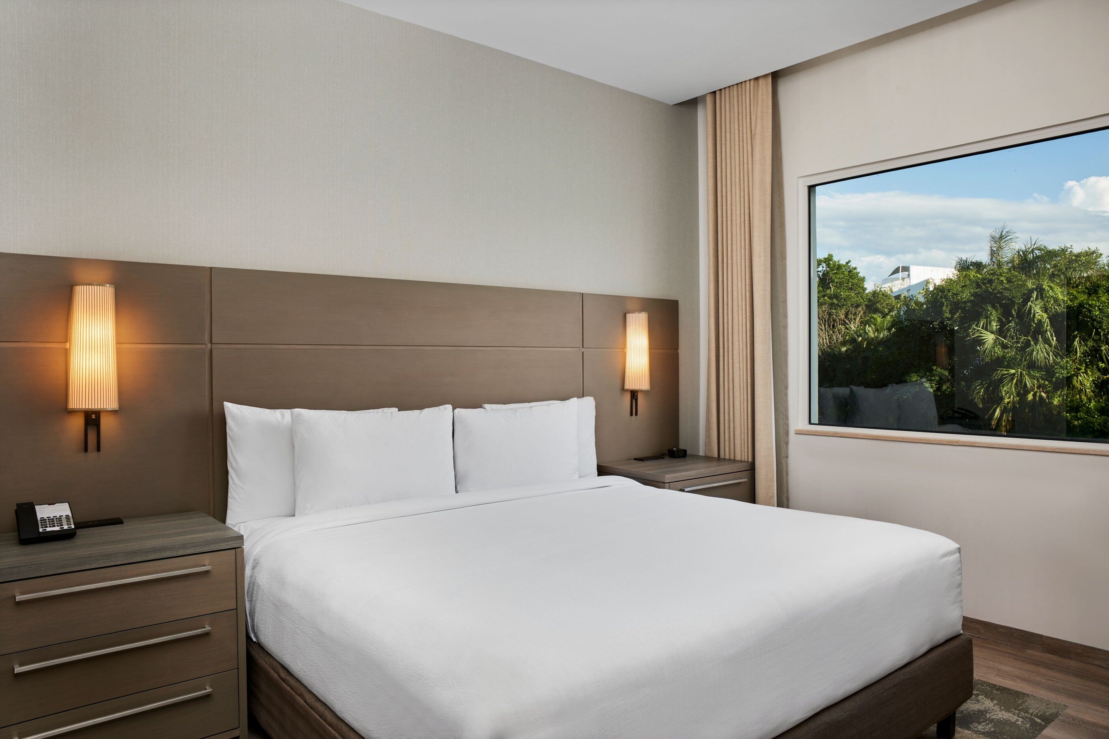 A modern hotel room with a large bed, two bedside tables, a telephone, and a window with a view of greenery.