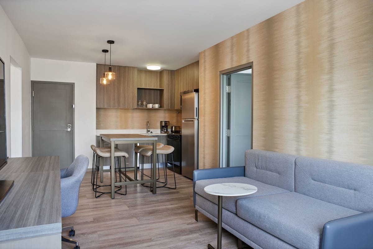Una cocina moderna con armarios de madera, un comedor con una mesa y taburetes de bar, y una sala de estar con un sofá y una pequeña mesa redonda.