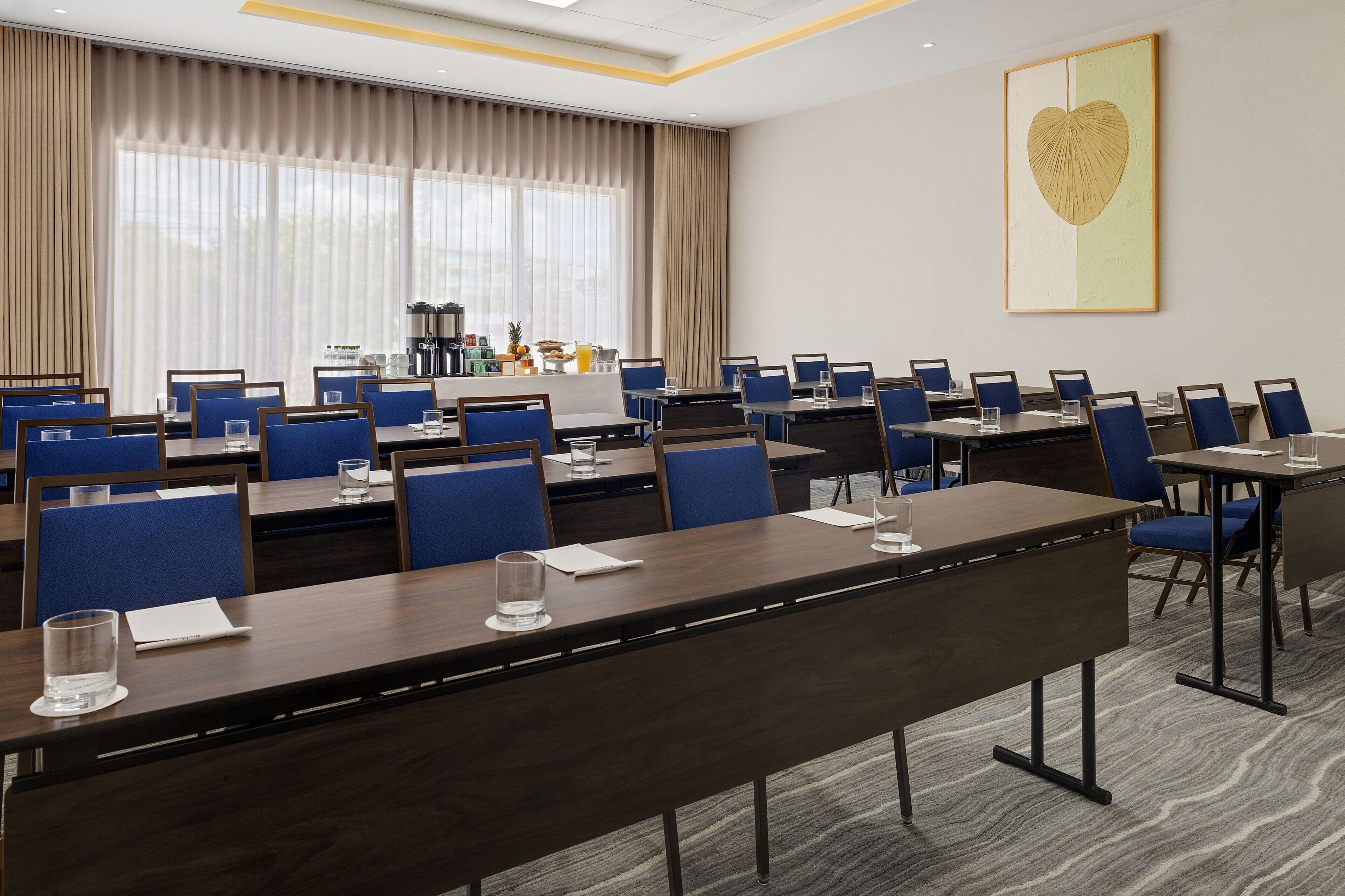 A conference room with multiple tables and chairs, a coffee station, and a large window with curtains.