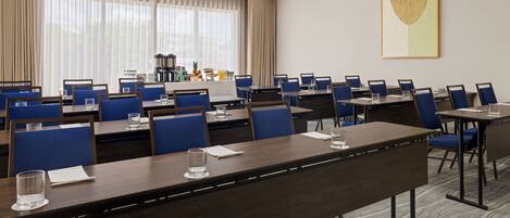 A conference room with multiple tables and chairs, a coffee station, and a large window with curtains.