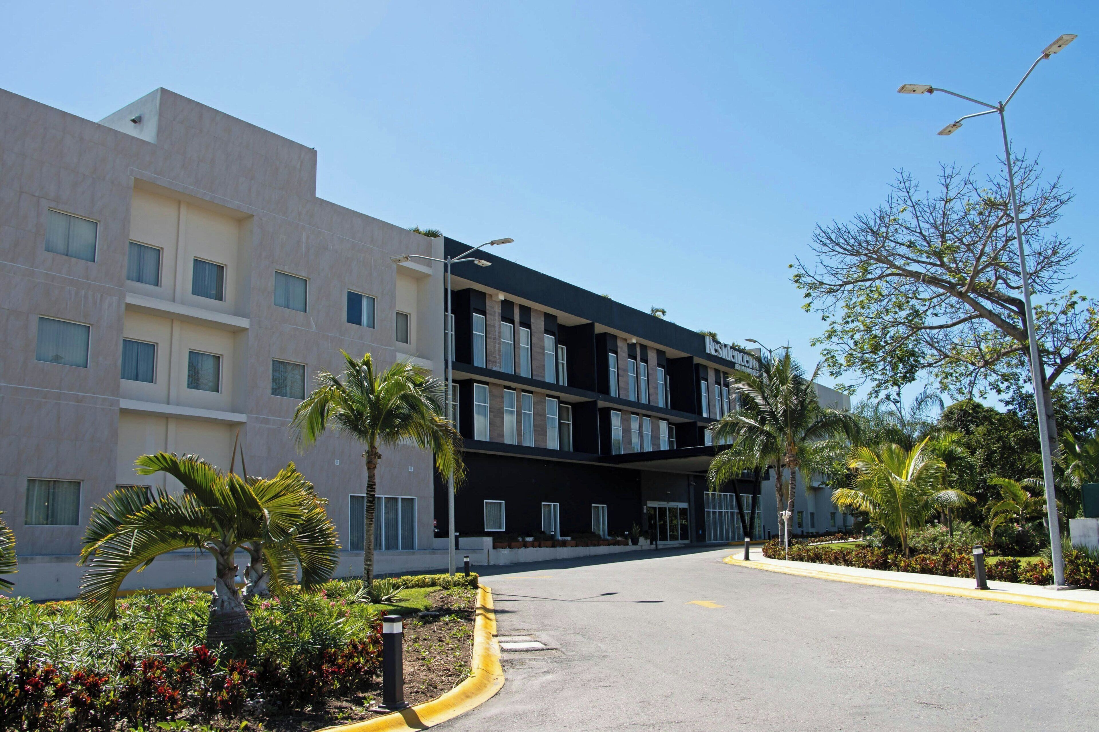 Un hotel moderno con una estructura de dos pisos, un acceso cubierto y un área ajardinada con palmeras y arbustos.