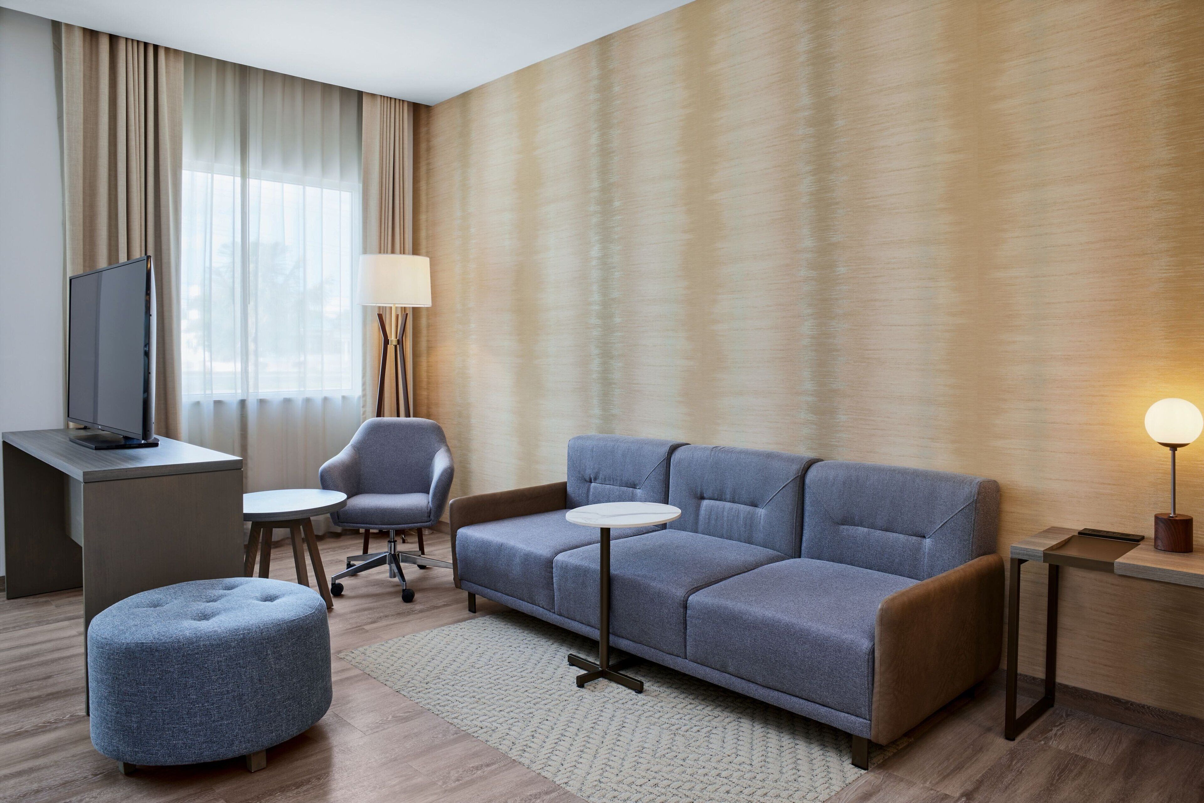 Et moderne hotelværelse med en grå sofa, en rund skammel, et skrivebord med et fladskærms-tv, et lille rundt bord og en gulvlampe.