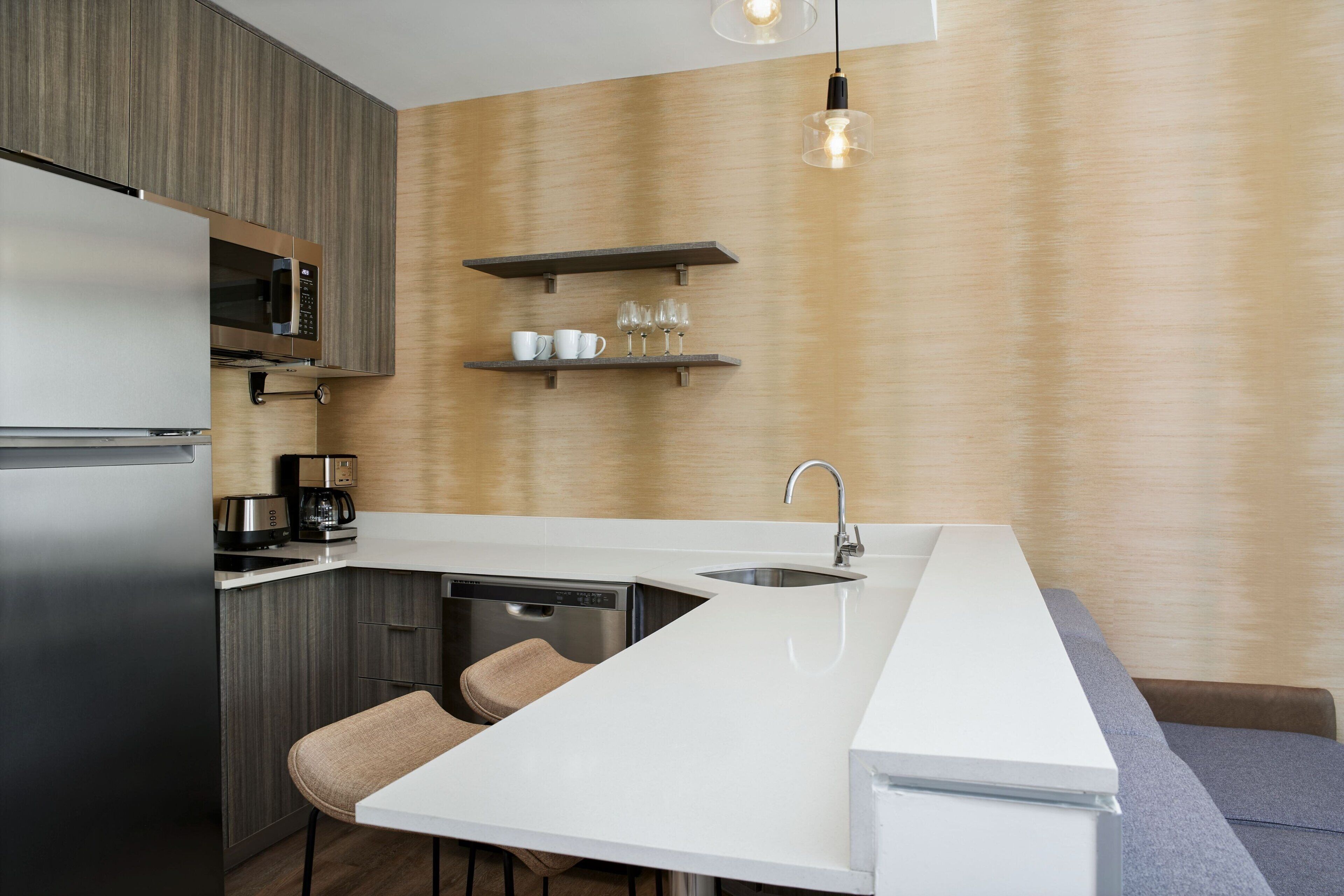 A modern kitchen with a built-in microwave, coffee maker, and dishwasher. There are two bar stools and a white countertop.