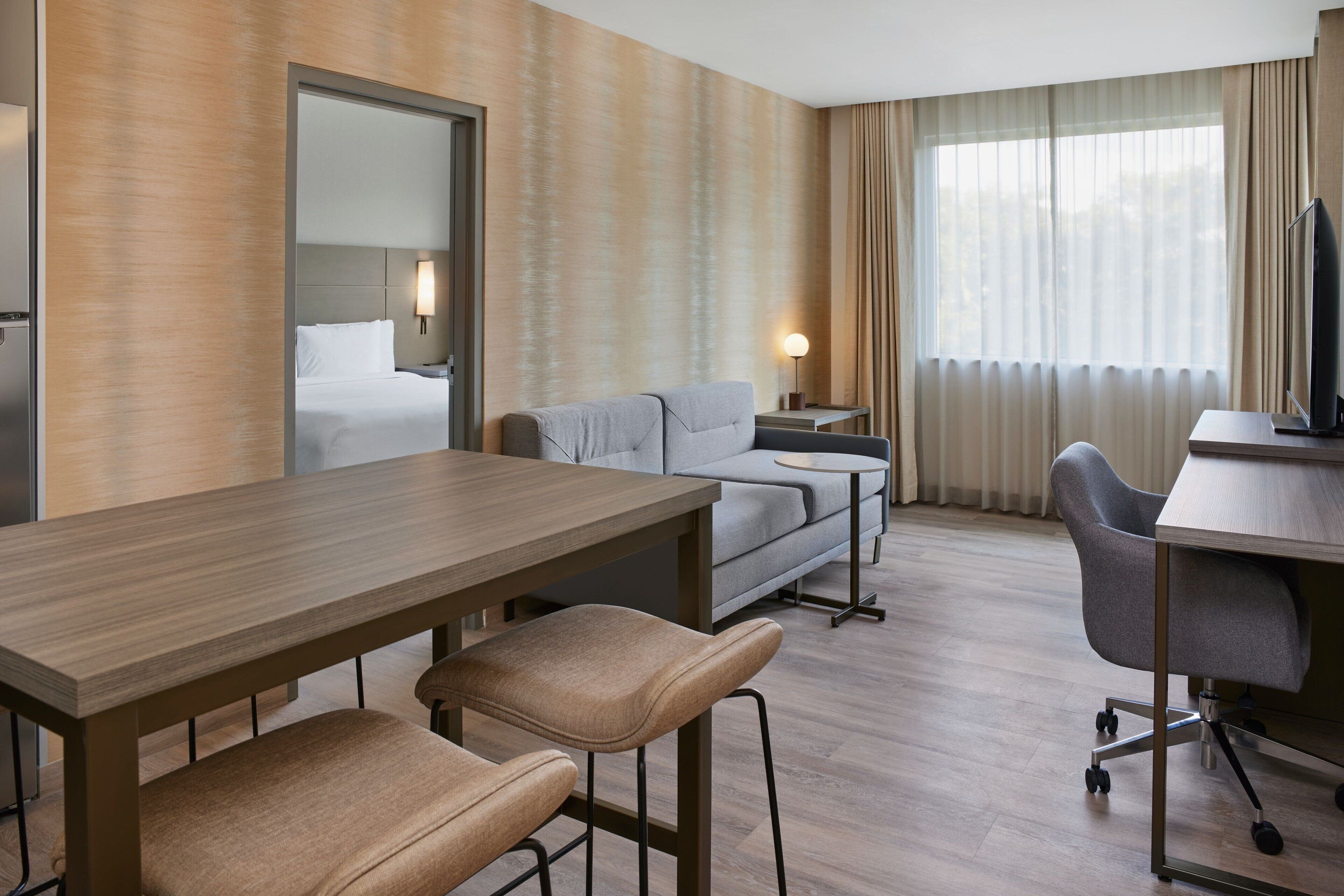 Habitación de hotel con escritorio de madera, dos sillones cómodos, un sofá, televisor, mesita de noche con lámpara y un ventanal grande con cortinas.