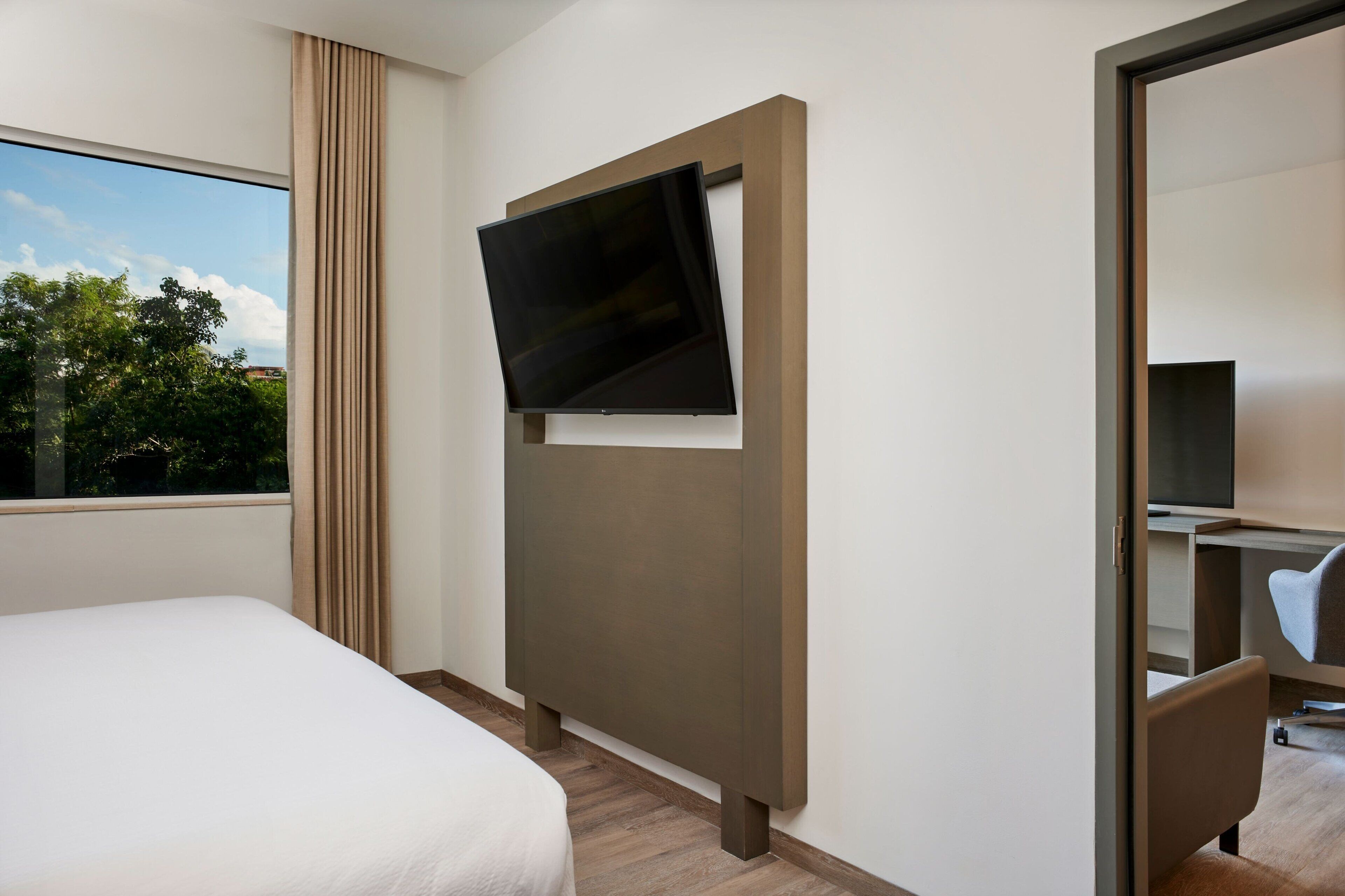 A hotel room with a bed, a TV mounted on a wall, a desk with a chair, and a large window with curtains.