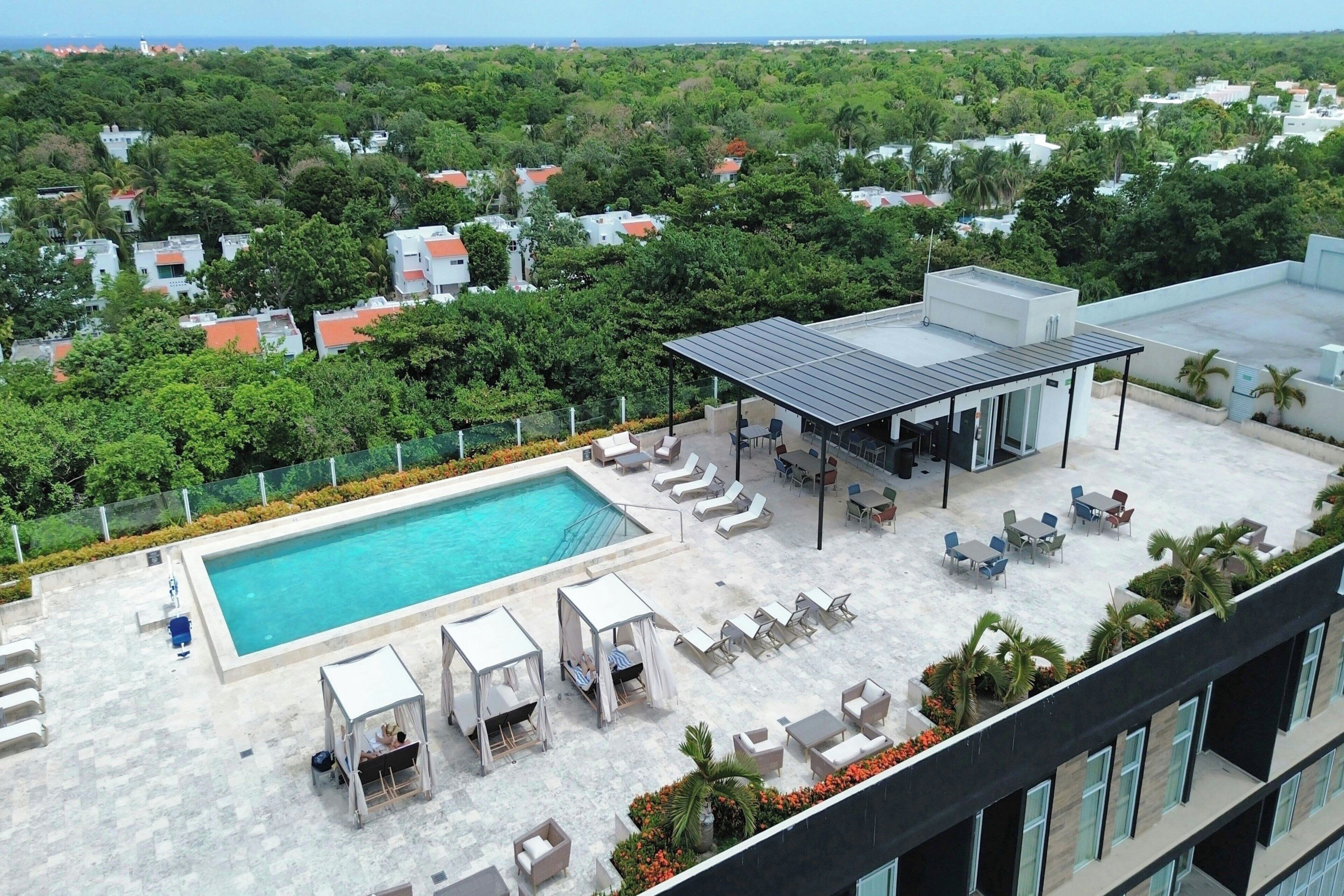 A rooftop pool area with a swimming pool, sun loungers, a covered seating area, and a view of surrounding greenery and buildings.