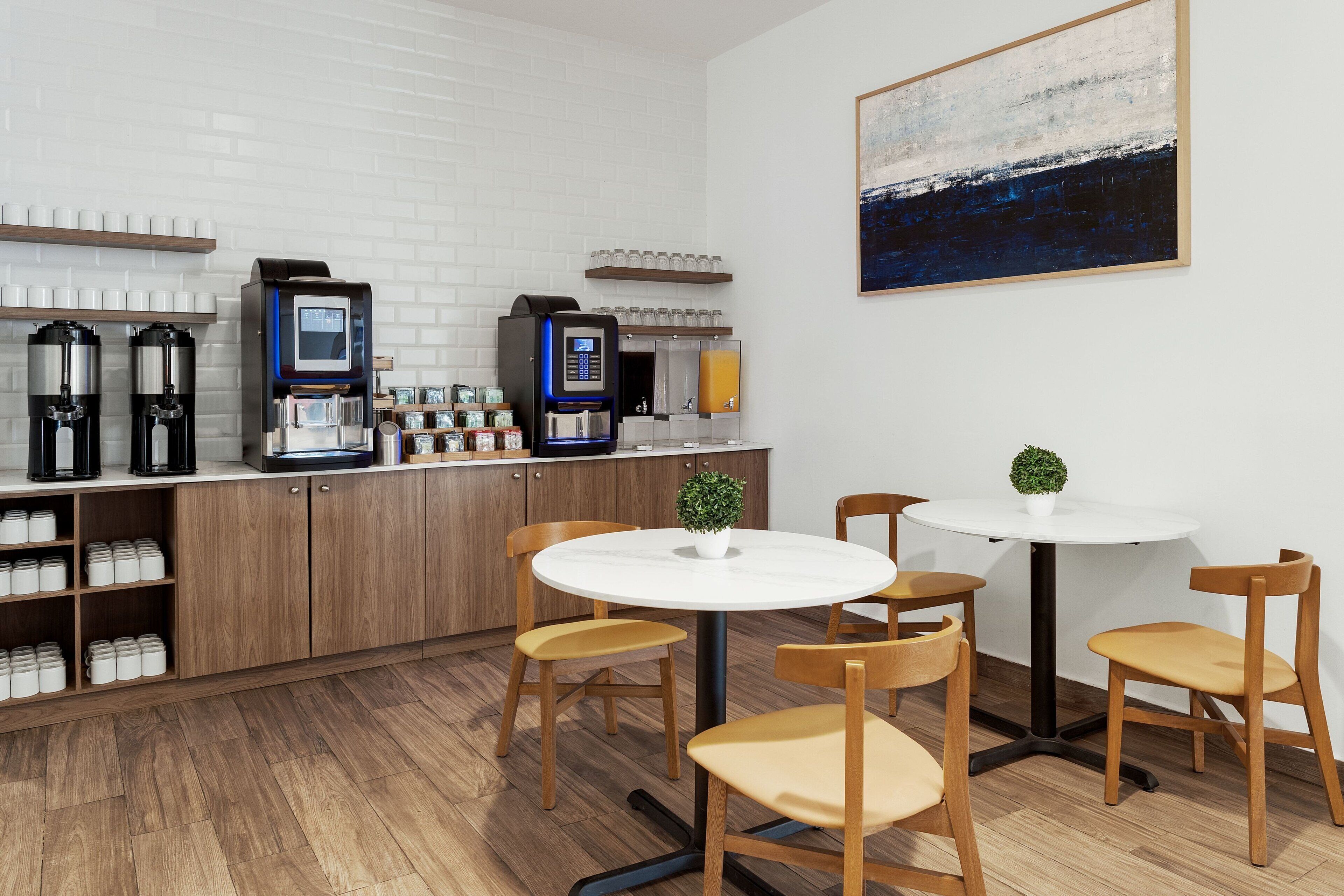 A modern kitchen with coffee machines, a white brick wall, and a painting. There are wooden chairs and a small table with a plant.