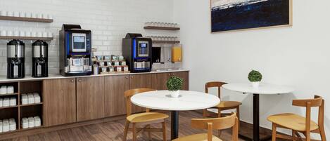 A modern kitchen with coffee machines, a white brick wall, and a painting. There are wooden chairs and a small table with a plant.