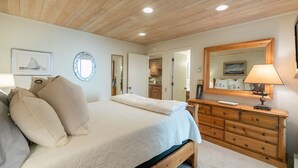 Un dormitorio con techo de madera, una cama con ropa de cama color beige, un aparador, un espejo y una lámpara.