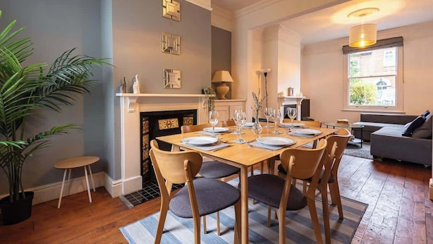 A dining area with a wooden table set for six, flanked by chairs with cushioned seats. A potted plant is visible on the left, and a fireplace with a mantel is on the right.