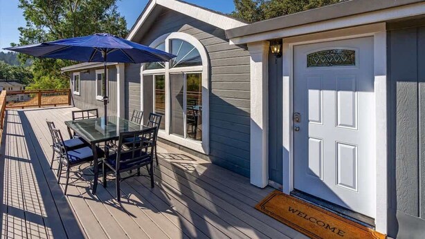 A house with a deck, a dining table with chairs, a blue umbrella, and a welcome mat. - Calistoga Vineyard Getaway - Hot Tub and Tastings Included (Calistoga)