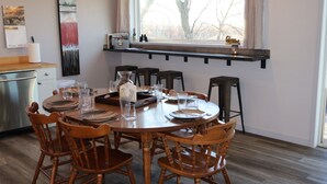 A dining area with a round wooden table set for six, surrounded by wooden chairs. A window with a view of trees is above the table.