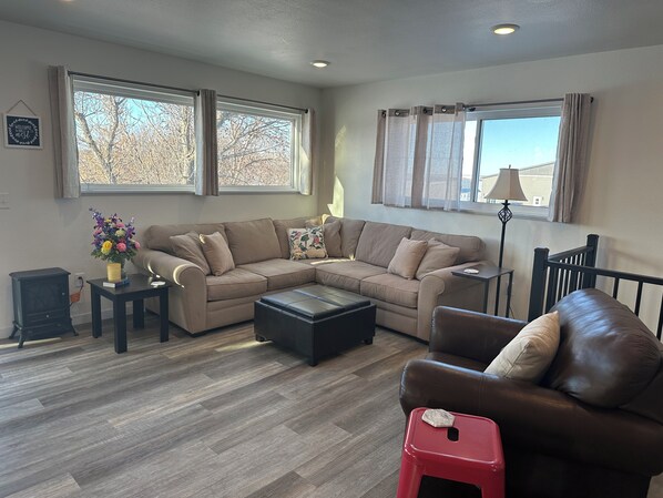 A living room with a sectional sofa, a coffee table, a red stool, and a lamp. There are two windows with curtains, a small table with a vase of flowers, and a wall-mounted picture frame.