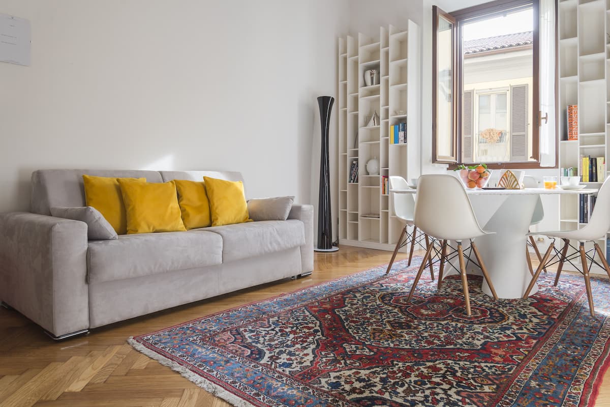 A living room with a grey sofa, yellow cushions, a patterned rug, a tall black vase, and a white dining area with chairs and a table.
