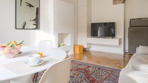 Una sala moderna con un sofá blanco, un televisor montado en una pared blanca, una mesa de comedor con fruta y una taza, y una alfombra estampada.