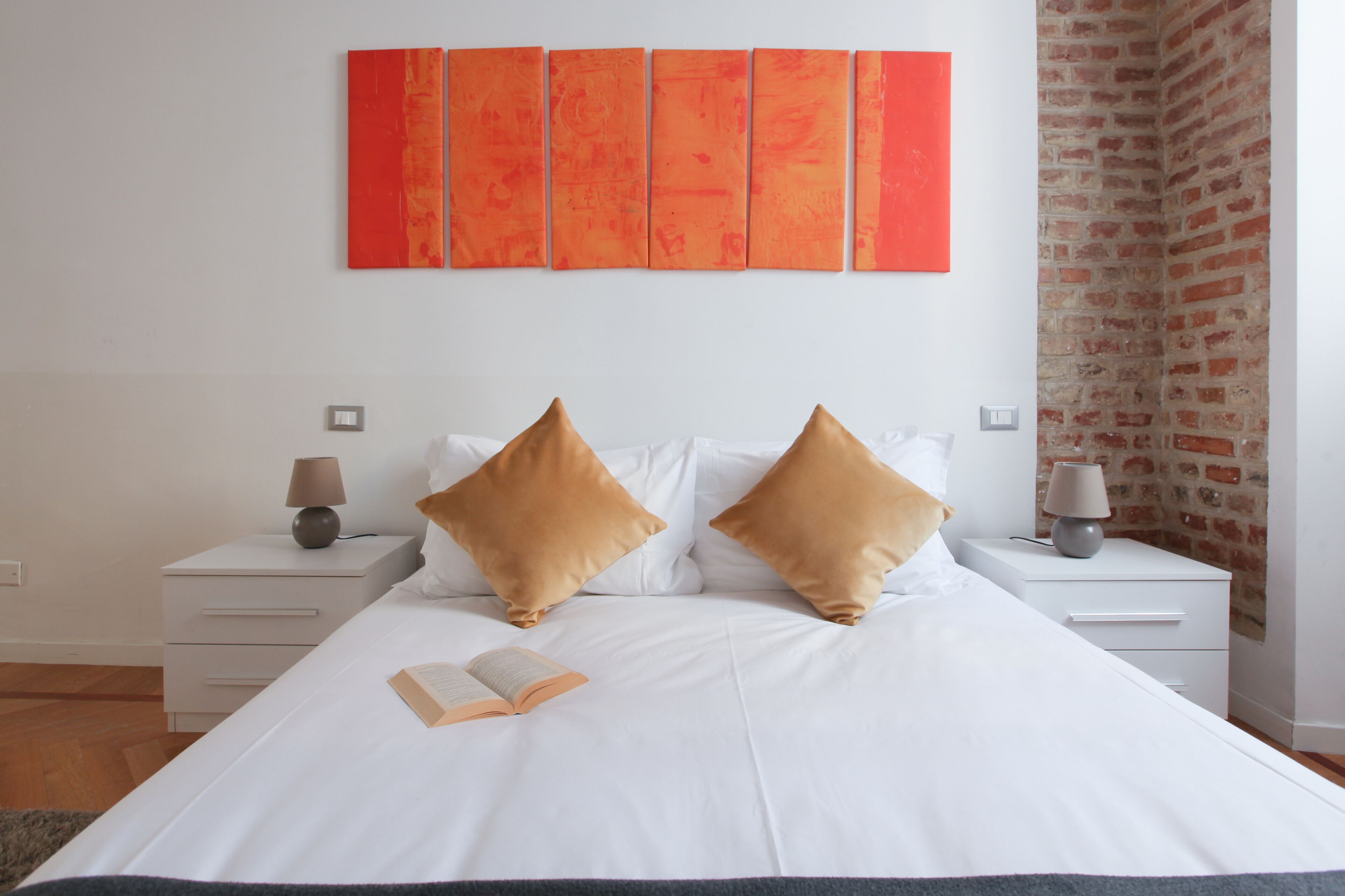 Una cama bien hecha con dos almohadas, una mesita de noche con lámpara y una pared con cinco paneles anaranjados.