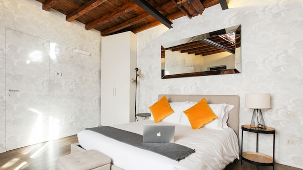 Un dormitorio moderno con techo de madera, una cama grande con ropa de cama blanca y almohadas naranjas, un ordenador portátil sobre la cama, una mesita de noche con una lámpara y un pequeño banco.